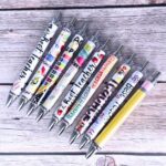 A close-up view of ten YJ PREMIUMS multicolor retractable ballpoint pens with various teaching-themed designs.